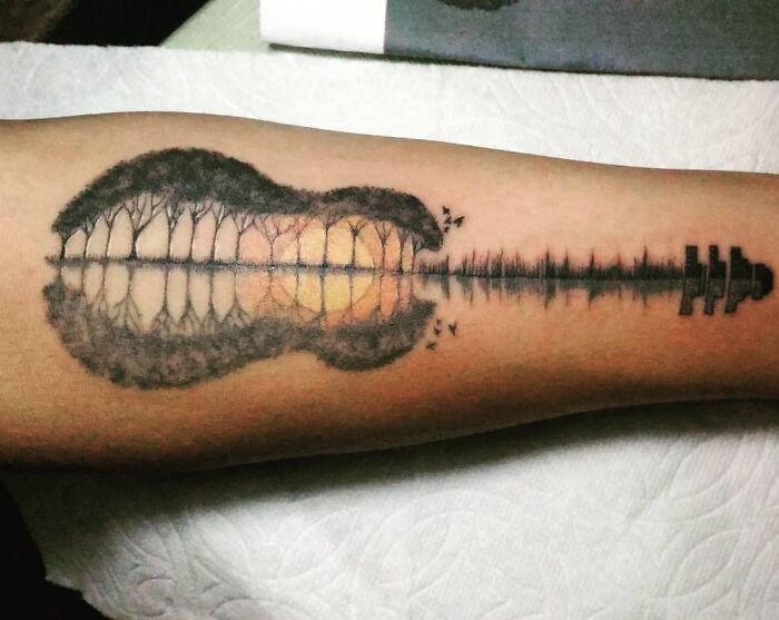 Optical illusion tattoo of a guitar silhouette with a landscape reflection on an arm.