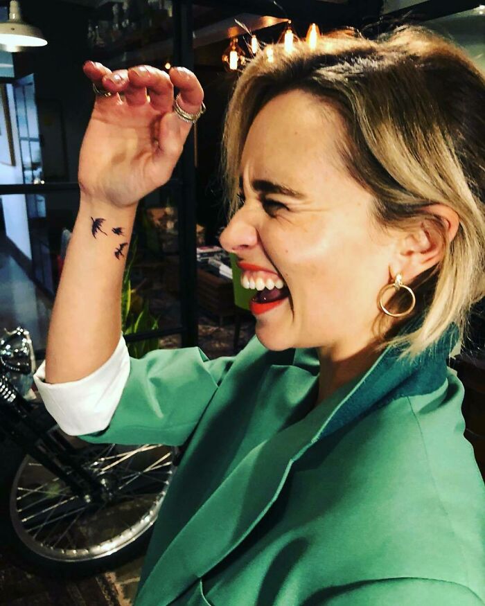 Emilia Clarke, smiling with bird tattoos on wrist, wearing a green jacket in a cozy setting.