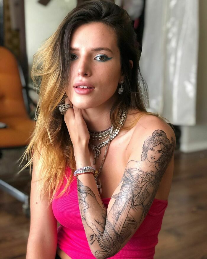 Woman in a pink top showing detailed celebrity tattoo on her upper arm in a casual indoor setting.