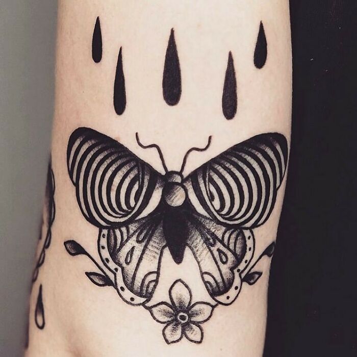 Optical illusion tattoo of a butterfly with intricate patterns and teardrop shapes above it on skin.