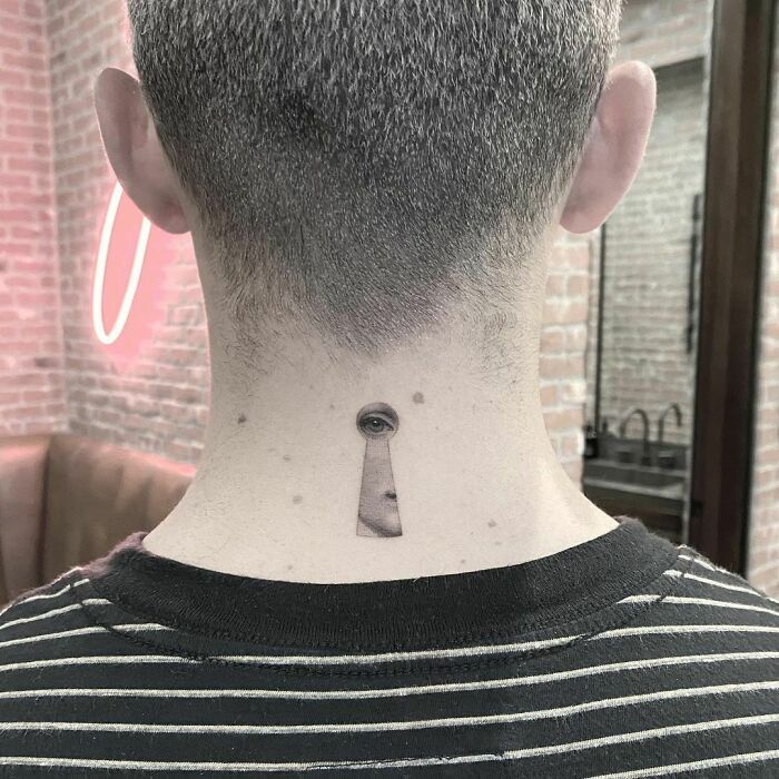 Celebrity tattoo of a keyhole with an eye on the back of a neck, in a brick-walled room with striped shirt visible.