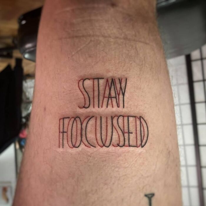 Optical illusion tattoo with distorted text "STAY FOCUSED" on skin, creating a mind-bending visual effect.