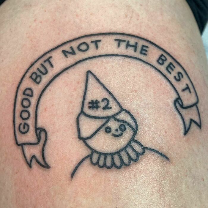 Funny tattoo of a smiling cartoon face with #2 cap, banner reading "Good but not the best" above.