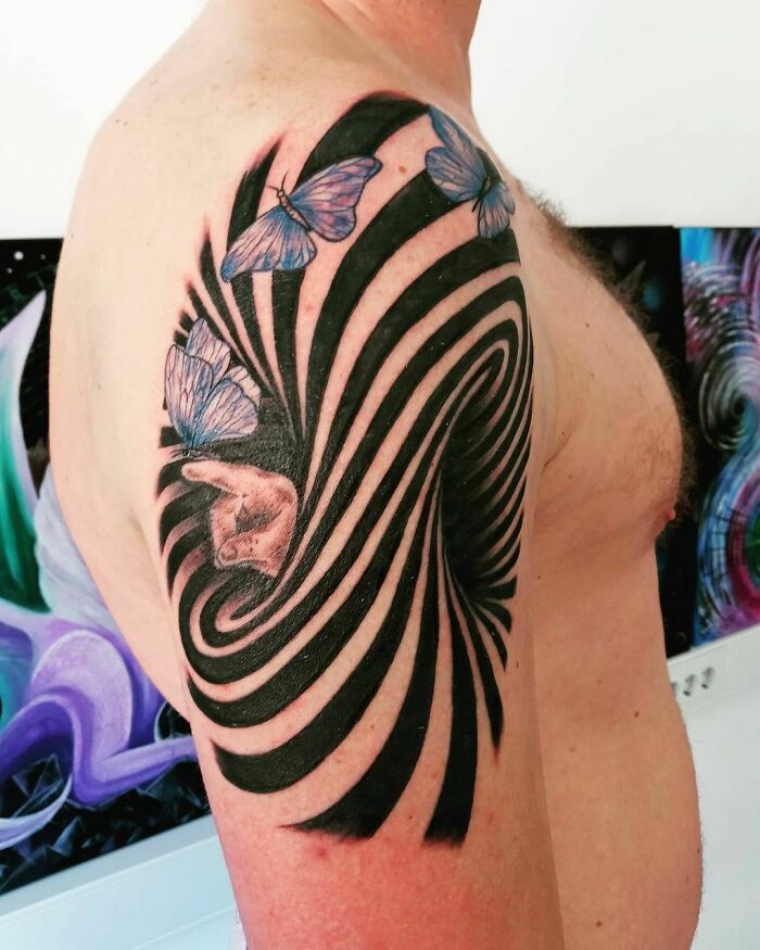 Optical illusion tattoo design with swirling black patterns and blue butterflies on a person's upper arm.