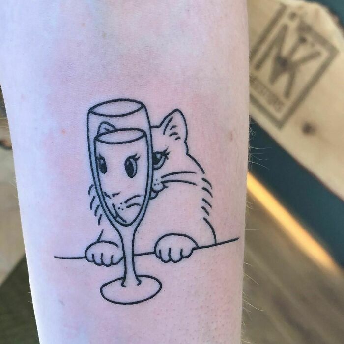 Funny tattoo of a cat humorously aligned with a wine glass on an arm.