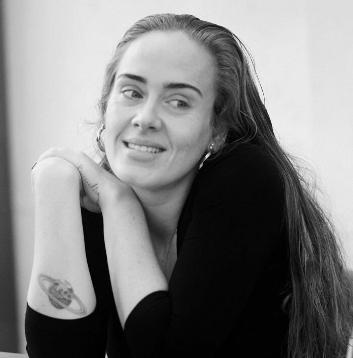 Person with long hair in a black shirt showing a planet tattoo on their arm, smiling in a relaxed setting.
