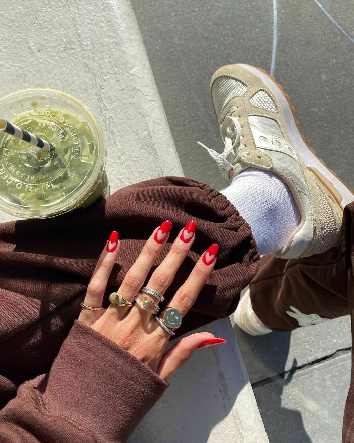 Stylish red nail art with heart tips as seen with layered rings, brown outfit, and casual sneakers.
