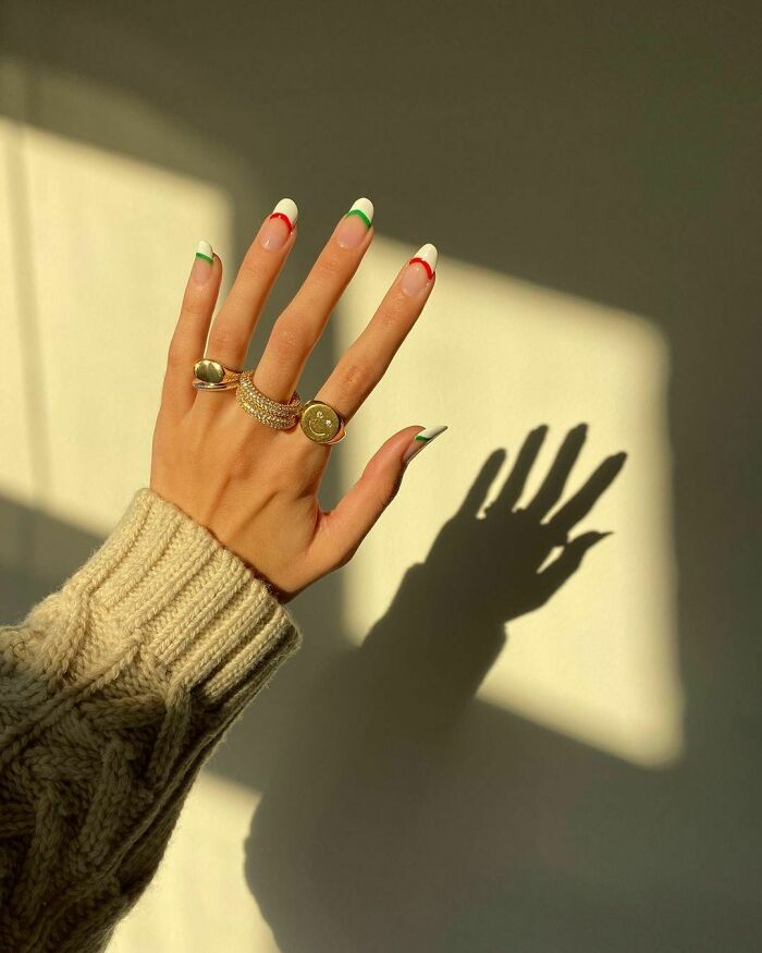Hand with colorful nail art ideas featuring red and green tips, wearing gold rings, casting a shadow on a beige wall.