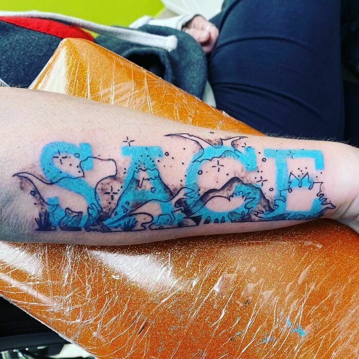 An optical illusion tattoo of dinosaurs creatively integrated within blue letters on a forearm.