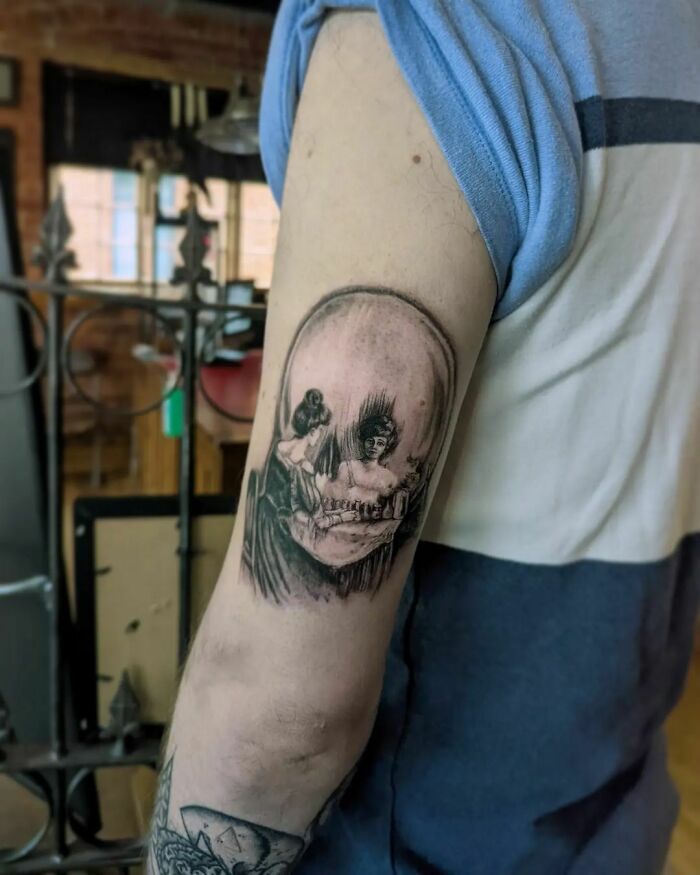 Optical illusion tattoo on arm, depicting a skull that blends into a scene with two figures.
