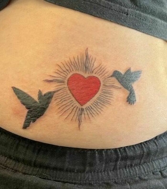 Tattoo of a red heart with rays, flanked by two black birds, on skin.