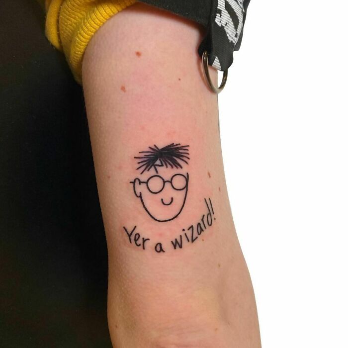 Funny tattoo of a simple cartoon face with glasses and text "Yer a wizard!" on an arm.