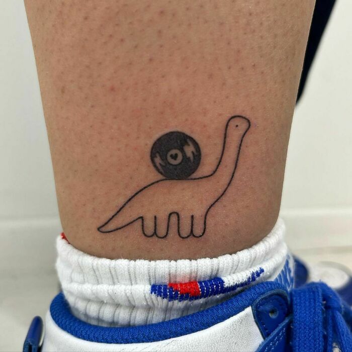 Funny tattoo of a dinosaur carrying a vinyl record on its back, located above an ankle wearing a white sock and blue shoe.