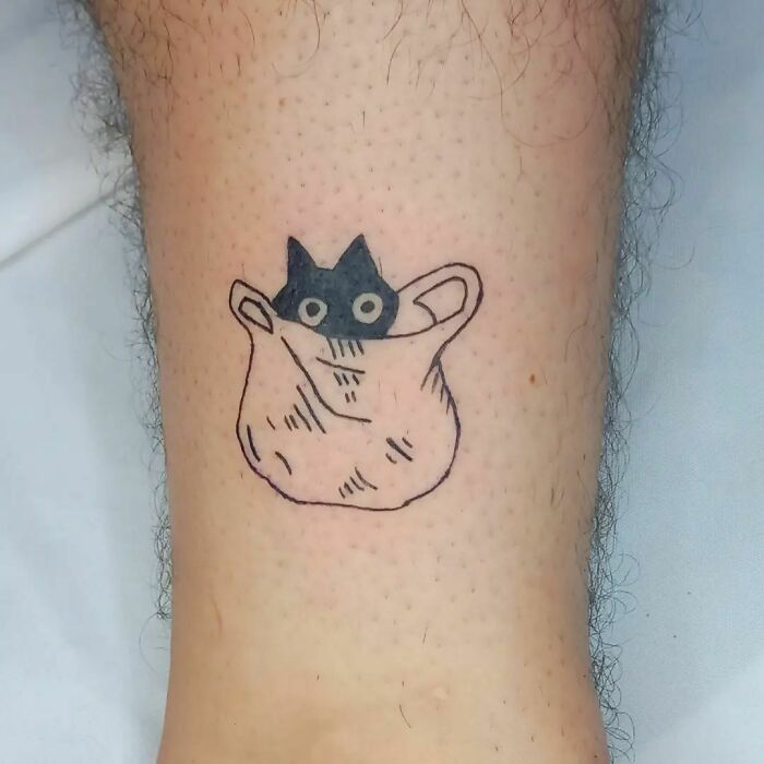 Funny tattoo of a cartoon cat peeking out from a bag on a person's leg.