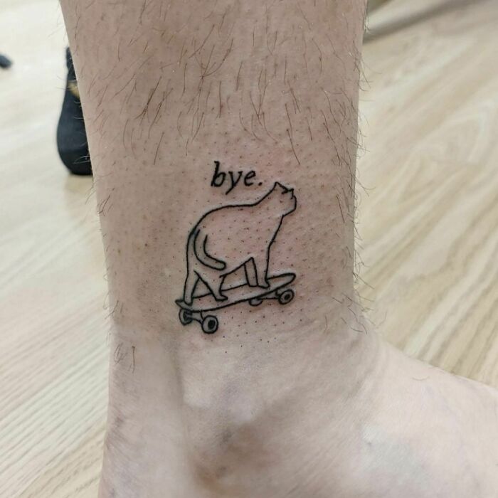 Funny tattoo of a cat on a skateboard with the word "bye" on an ankle.