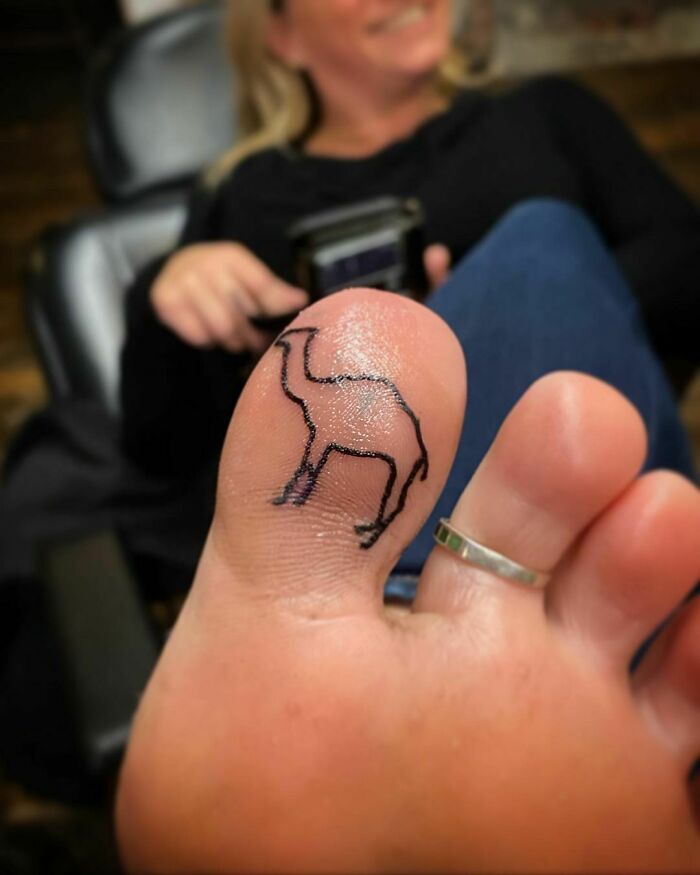 A funny camel tattoo on a person's big toe with a woman smiling in the background.