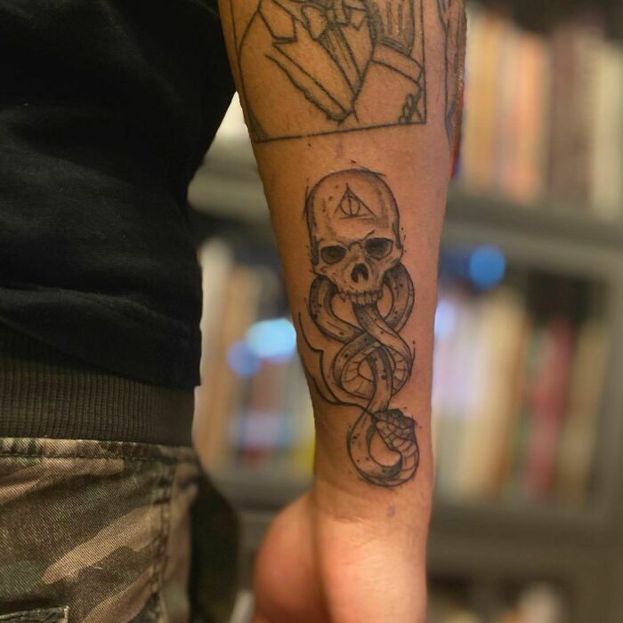 Arm with intricate Harry Potter tattoo featuring a skull and snake, symbolizing a dark wizarding theme, in a library setting.