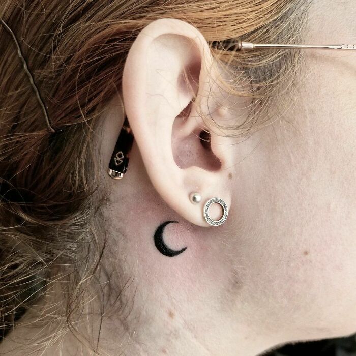 A minimal tattoo of a crescent moon behind the ear with piercings visible.