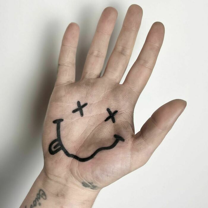 Funny tattoo of a crazy face drawn on the palm, featuring a playful smile and crossed-out eyes.