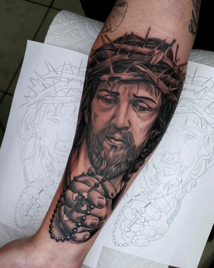 Tattoo of Jesus with crown of thorns on forearm, over sketched designs, focusing on attractiveness perception.