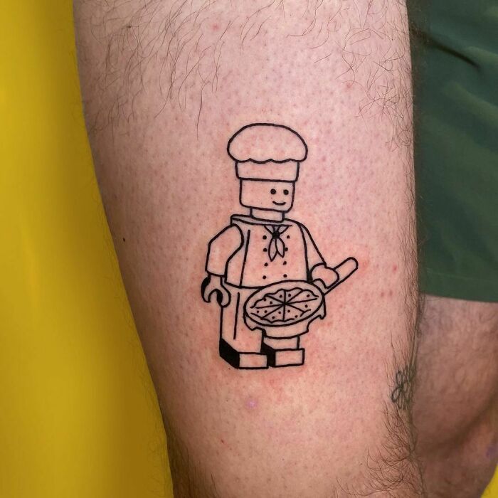 Funny tattoo of a Lego man chef holding a pizza on a leg, adding a playful touch.