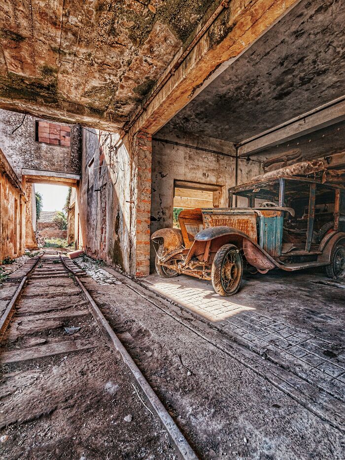 Rusty classic car parked inside an abandoned factory with old railway tracks leading outside in a dilapidated setting.