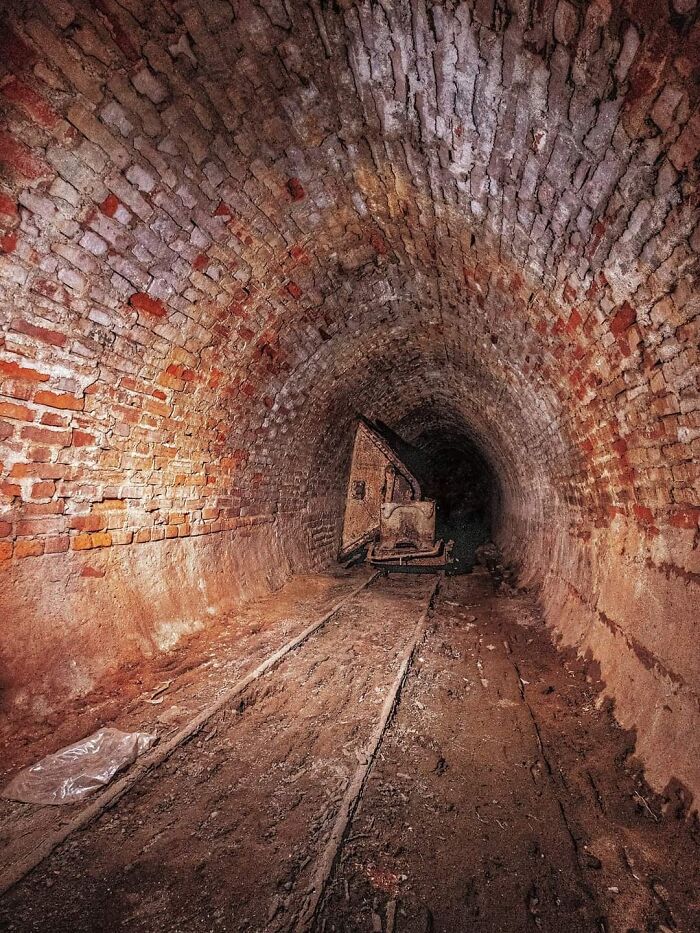 Abandoned factory tunnel with brick walls and old rusty cart on narrow tracks during urban exploration.