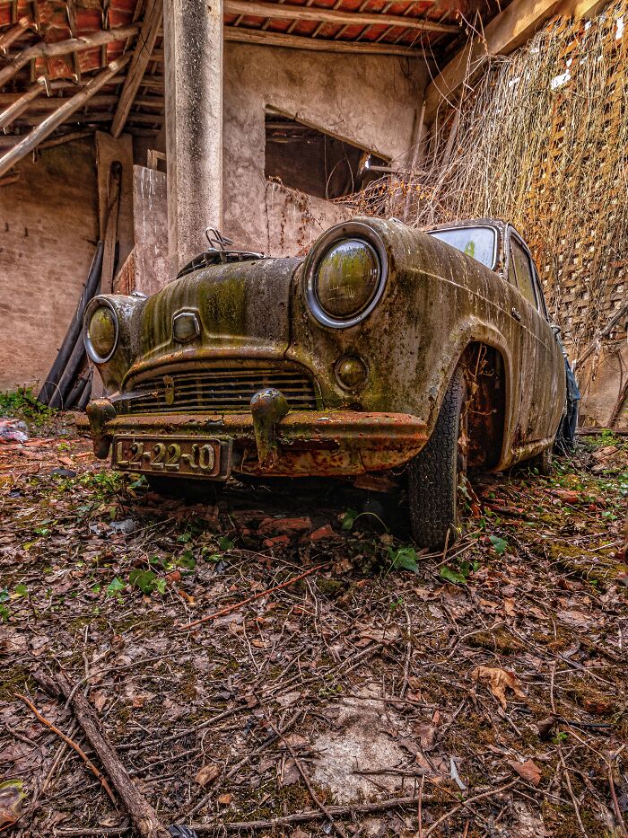 Rusty classic car covered in moss and grime inside an abandoned factory with overgrown debris on the ground.