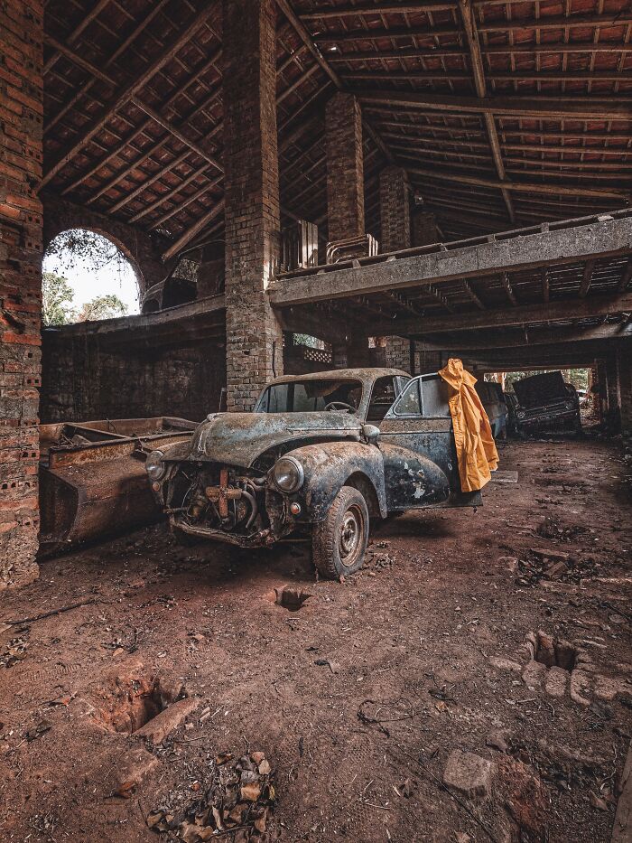 Rusty classic car abandoned inside an old factory with exposed brick pillars and a dirt-covered floor surrounding it.