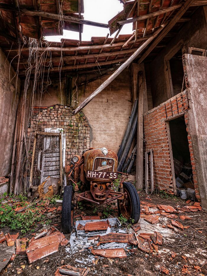 Rusty classic car abandoned inside a dilapidated factory with broken roof tiles and overgrown vegetation.
