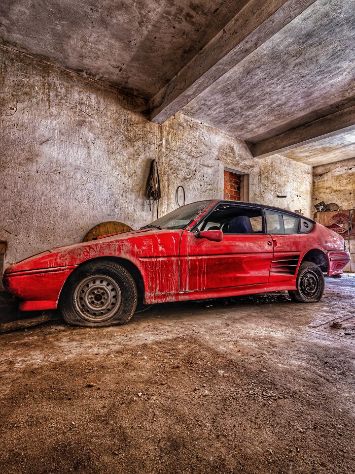 Abandoned factory interior with a dusty classic red car showing signs of wear and flat tires on a concrete floor.