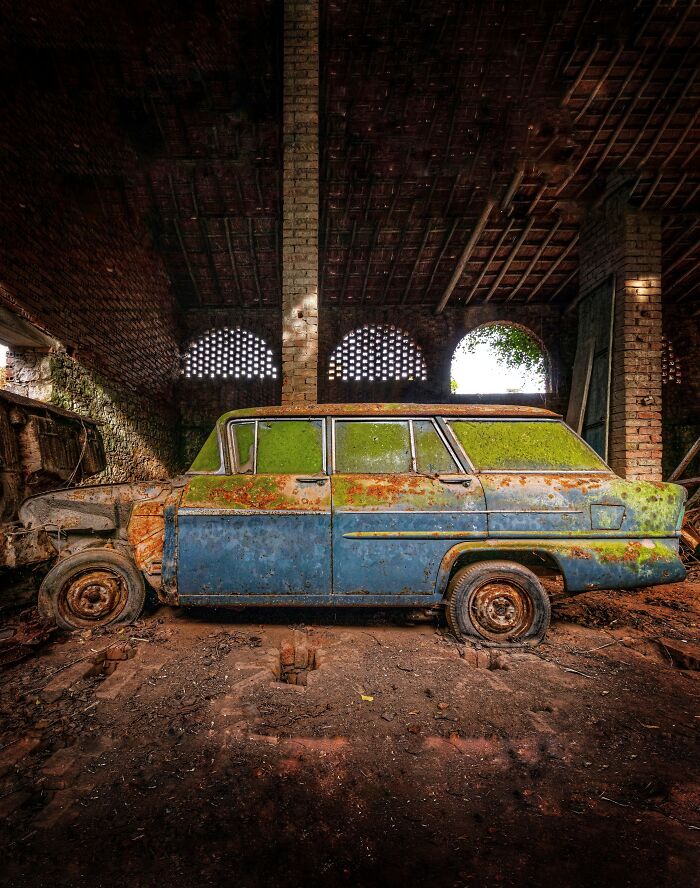 Rusty classic car covered in moss, abandoned inside a deteriorating factory with brick walls and arched windows.