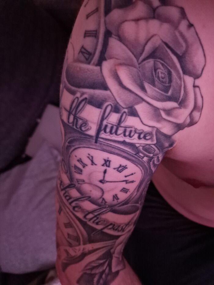 Arm tattoo featuring a clock, rose, and the phrase "the future holds the past" in detailed black and gray ink.