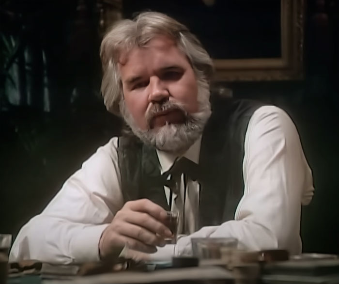 Kenny Rogers wearing a black vest, sitting at a table, representing famous people who got married multiple times.