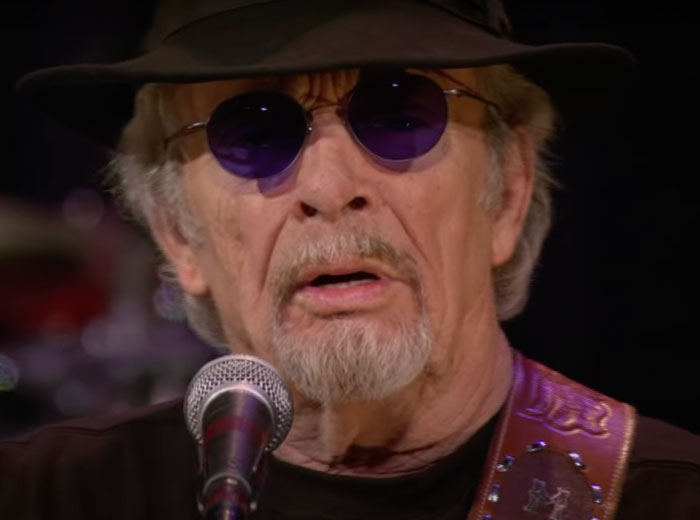 Merle Haggard wearing glasses and hat, singing into microphone, representing famous people who got married multiple times.