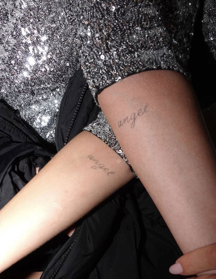 Two arms showing matching "angel" tattoos in elegant script, against a backdrop of glittery fabric, highlighting celebrity tattoos.