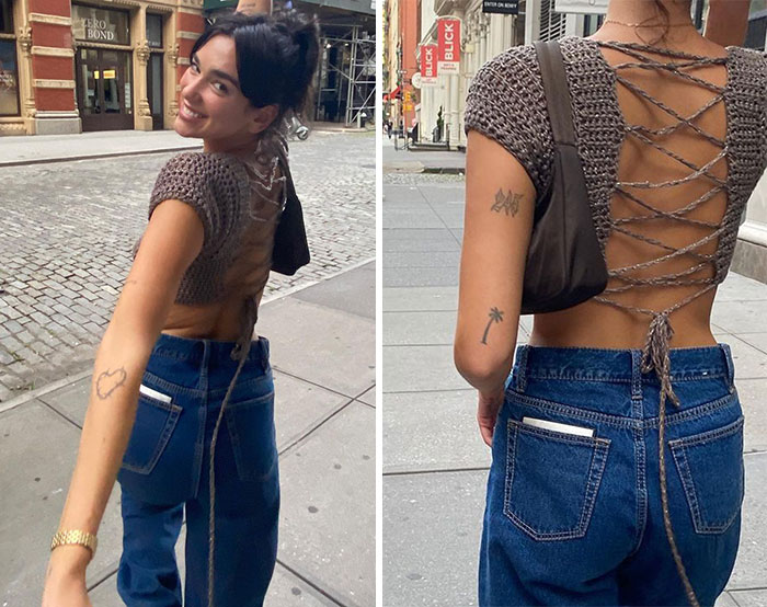 Person with celebrity tattoos on arm, wearing a crochet top and jeans, walking on a city street.
