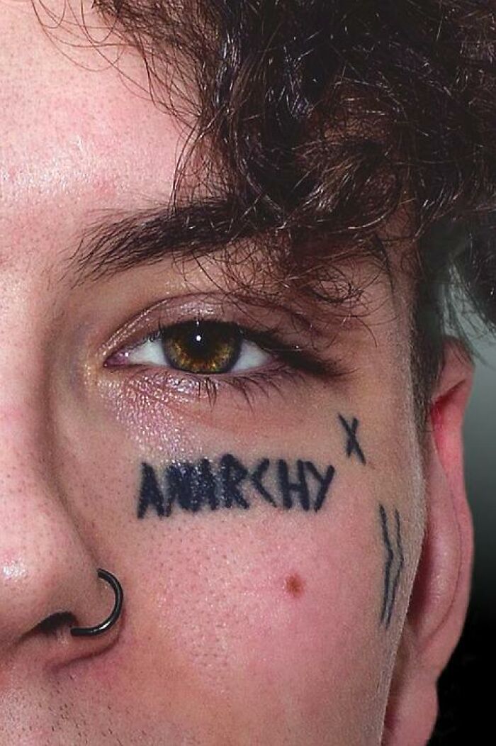 Facial tattoo of the word "anarchy" and symbols under an eye, with a nose piercing, reflecting a bold style choice.