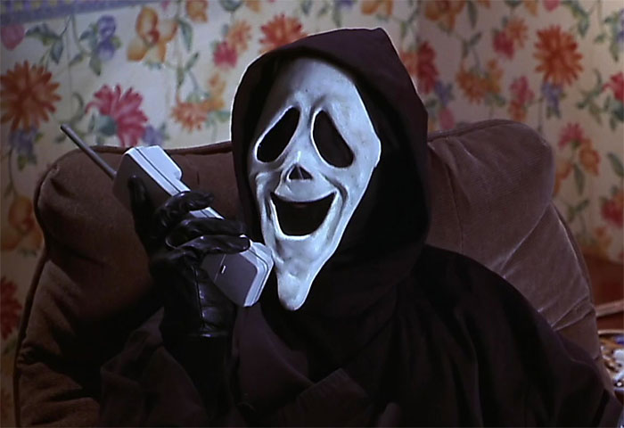 Person in a black cloak wearing a ghost face mask while holding a vintage mobile phone, representing big trends from the past.