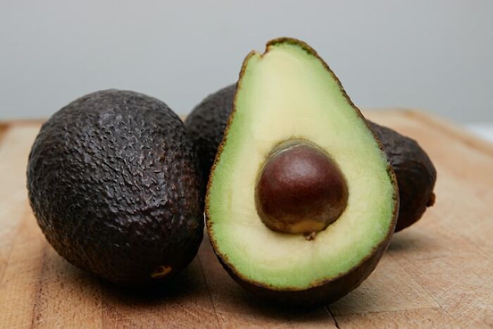 Three avocados on a wooden surface, with one cut open showing the seed, highlighting peasant food affordability issues.
