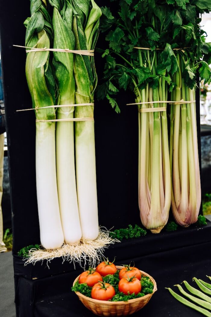 Bundles of fresh leeks, celery stalks, and a basket of ripe tomatoes displayed at a market stall.