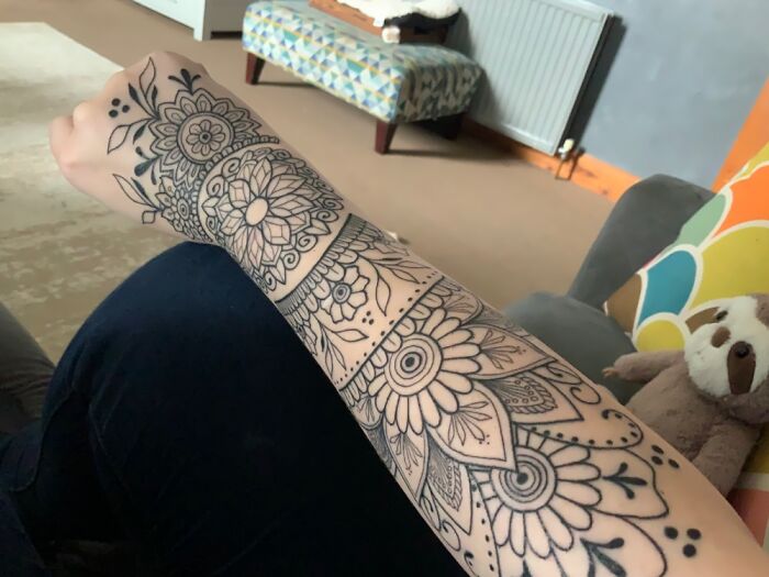 Arm with intricate tattoo featuring floral and geometric designs in a cozy living room setting.