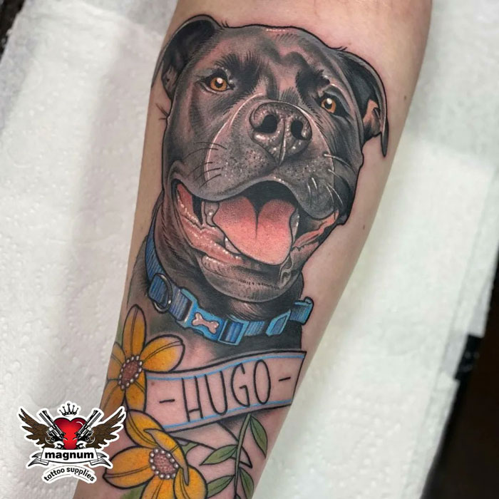 Adorable pet tattoo of a dog with a blue collar and name "Hugo," surrounded by yellow flowers on a forearm.