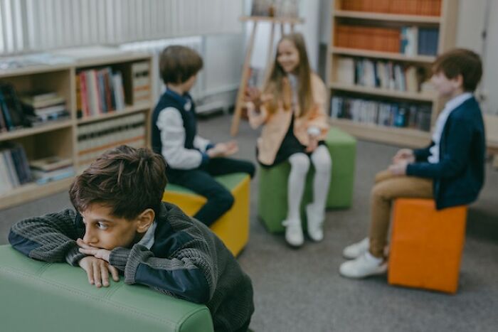 A pensive child leaning on a couch while other kids talk in a library setting representing bad parent concerns.