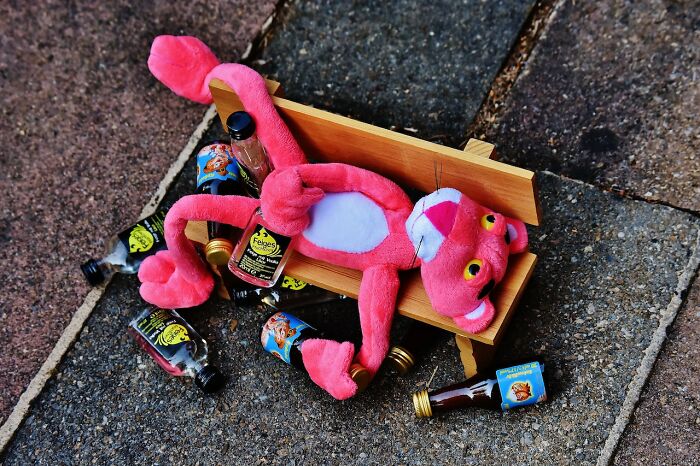 Pink plush toy lounging on a small bench surrounded by empty miniature bottles showing unintended bad outcomes.
