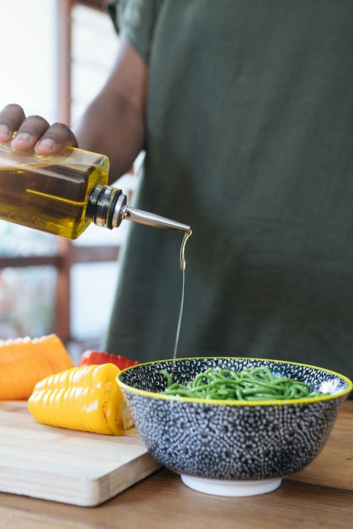 Person pouring oil into a bowl of green pasta with sliced vegetables on a wooden cutting board at home.