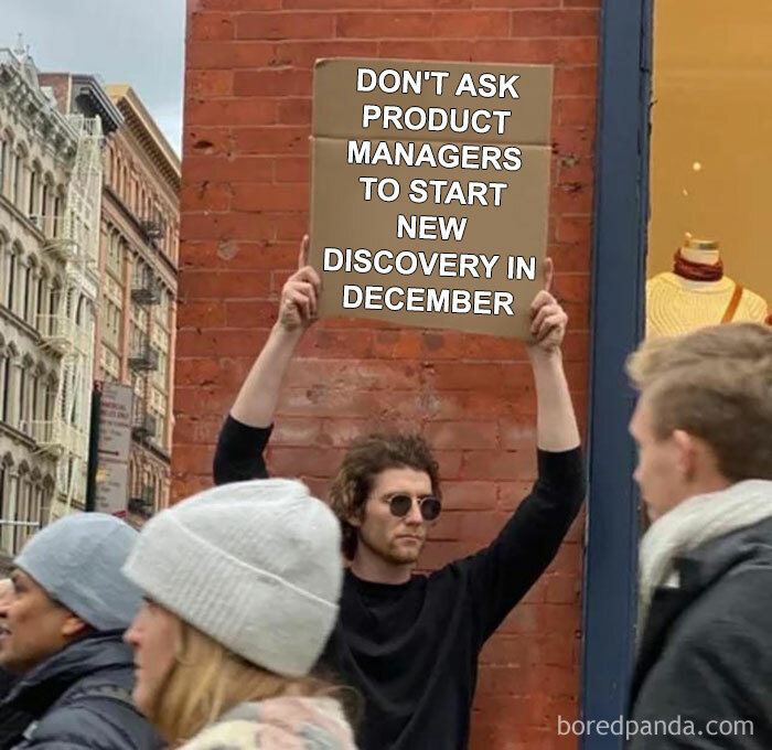 Man holding sign about product manager problems in December, standing against a brick wall in a busy urban area.