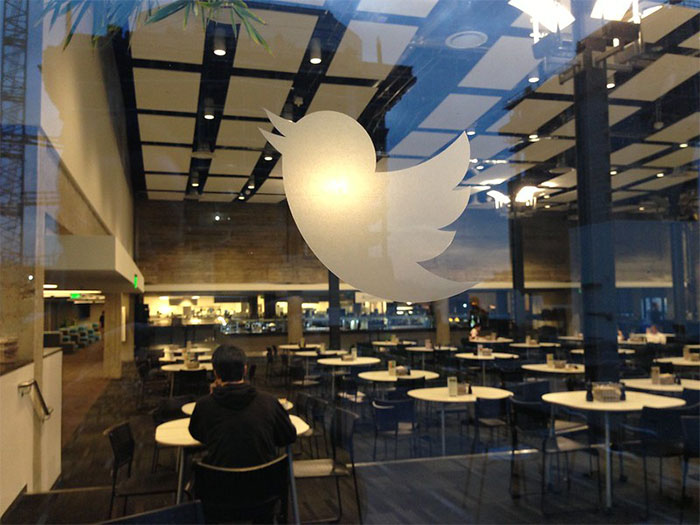 View through a window with a Twitter logo, showing a nearly empty modern cafeteria with few people inside.