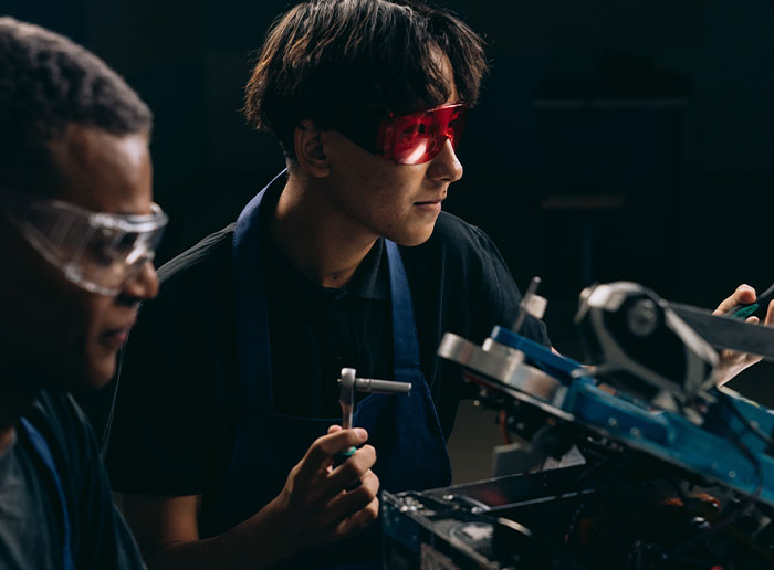 Two young technicians wearing safety glasses working carefully on electronic equipment in a dark workshop setting