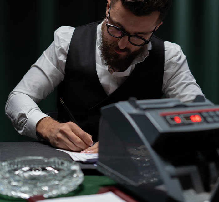 Man in glasses and vest writing by hand at desk with a vintage adding machine, illustrating professions that may soon disappear.